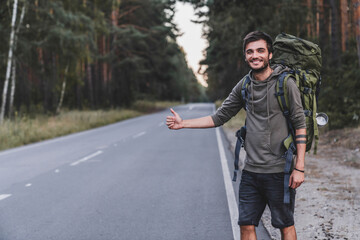 Caucasian young male traveler try to catch car on forest road. World travel road trip, holiday hitch hike concept. Handsome explorer traveling in solo trip alone, escaping from city life