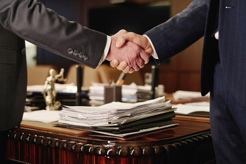 Closeup of two unrecognizable businessmen shaking hands standing next to table with pile of papers - obrazy, fototapety, plakaty