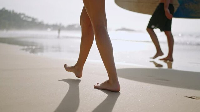People with surfboards go on wet sand, low angle back view. Surfers couple barefoot feet walk on wet sandy beach by sea surf. Footprints of man, woman going surfing on ocean waves on tropical island.