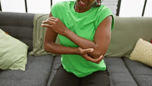 A mature black woman in a green shirt feels elbow pain while sitting on a gray couch indoors, expressing discomfort.