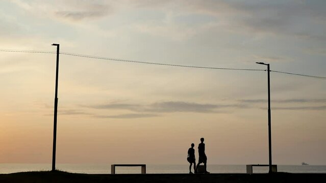 Silhouettes of a young man and woman leisurely walking together along the sea promenade in the evening at sunset. Slow motion of a married couple walking along the pier