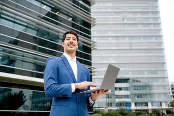 Fototapeta premium In a stance of readiness, a Hispanic business professional holding a laptop outdoors, with a glass building behind him, epitomizing the flexibility and mobility of modern business practices.