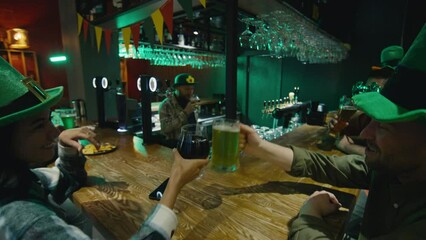 POV of clanking glasses of green and craft beer sitting with friends in green hats at bar counter on Saint Patricks day and celebrating with bartender