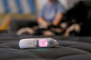 Close-up of a digital thermometer on a sofa, blurry background with a sick toddler kid sitting 
