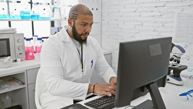 Focused black man in lab coat working on computer in modern laboratory setting
