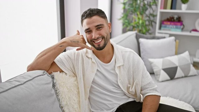 Cheerful young arabian man sitting on home sofa, joyfully making phone gesture with hand and fingers, smiling amidst friendly communication concept