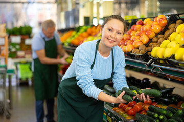 Positive middle-aged woman, skilled market assistant arranging organic cucumber on shelves in...