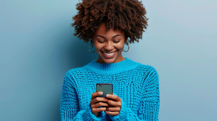 young woman with a vibrant afro hairstyle is smiling and looking at her smartphone, wearing a cozy turquoise turtleneck sweater against a matching turquoise background - obrazy, fototapety, plakaty