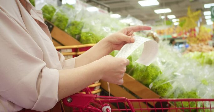 Woman against background of vegetables checks paper check after shopping for groceries at mall by checking Dear Amount bill in grocery cart. Increase in food prices. Woman checking grocery store cart.