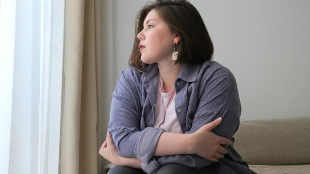 Contemplative young plus size woman seat beside window, her arms crossed, she looks outside, bathed in soft natural light.