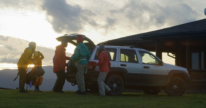 Full shot of diverse tourist friends arrive to rental house on hill. Happy hikers take luggage from trunk of car and talk. Hiking buddies during trip or expedition to mountains on their vacation.