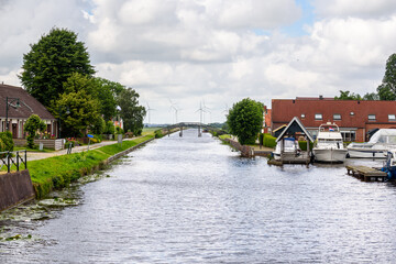 Canal through a small village in the countryside of Netherland on an overcast summer day. A wind farm is visible in background.