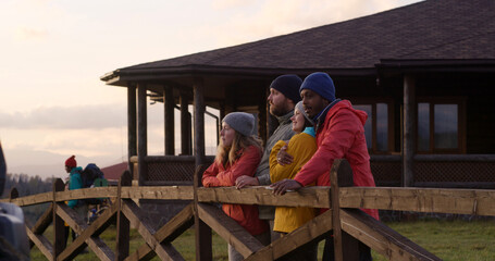 Group of multiethnic tourists stand near wooden fence, talk and look at beautiful sunset. Hiking friends relax during vacation trip in mountains. Stylish wooden cottage in the background. Slow motion.