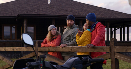 Group of multiethnic tourists stand near wooden fence, talk and look at beautiful sunset. Hiking friends relax during vacation trip in mountains. Stylish wooden cottage in the background. Slow motion.