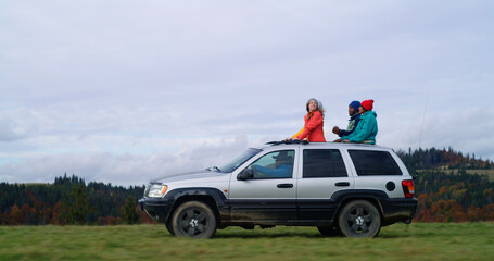 Group of diverse tourists together ride on vacation in mountains. Hikers standing up through sunroof of car sing, dance and wave hands on road trip. People enjoy drive in car hatch. Active recreation. - obrazy, fototapety, plakaty