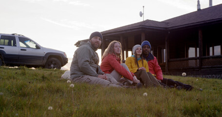 Group of multiethnic tourists sit on the grass at the hilltop and talk. Travelers or hiking friends enjoy nature during trip to the mountains. Stylish wooden cottage, car and dog in the background.