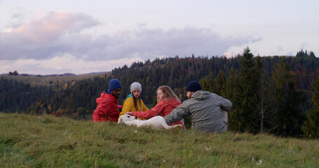 Fototapeta na wymiar Group of multiethnic tourists sit on the grass at the hilltop and pet a dog. Travelers or hiking friends enjoy nature during trip. Sunset and beautiful mountain landscape. Active recreation concept.