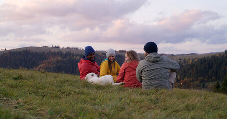 Group of multiethnic tourists sit on the grass at the hilltop and pet a dog. Travelers or hiking friends enjoy nature during trip. Sunset and beautiful mountain landscape. Active recreation concept.