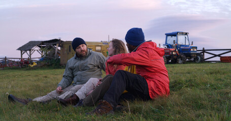 Diverse tourists or hiking buddies sit on the grass at the hilltop and pet dog. Group of travelers talk and enjoy nature during trip. Sunset and amazing mountain scenery. Outdoor exploration concept.
