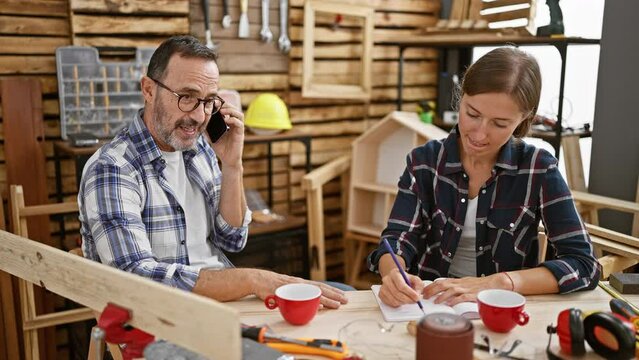 Two spirited carpenters, man and woman, partners in their booming carpentry business, cracking a high five, all while talking, taking notes on their smartphone amidst the woodwork indoors.