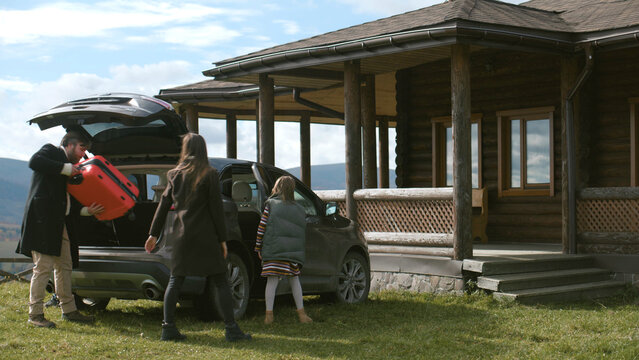 Happy Family Gets Out Of The Car, Walks With Suitcase To Modern House. Couple With Kids Settles Into Rented Wooden Cottage In Mountains For Vacation Or Moves To New Home Property. Real Estate Concept.