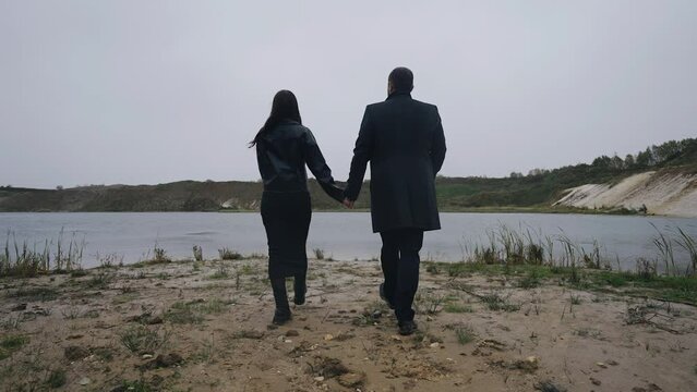 A man and a woman walk in the rain holding hands towards the shore of a sand quarry. The camera captures sadi in motion A very melancholic atmosphere