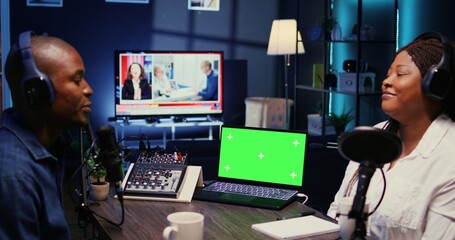 Green screen laptop next to vlogger show host and guest talking in neon lights ornate living room personal studio. Mockup notebook next to man and woman broadcasting discussion on online podcast