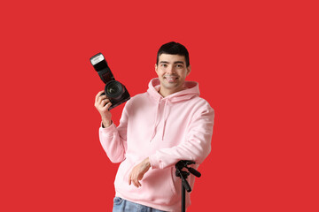 Young male photographer with professional camera and tripod on red background