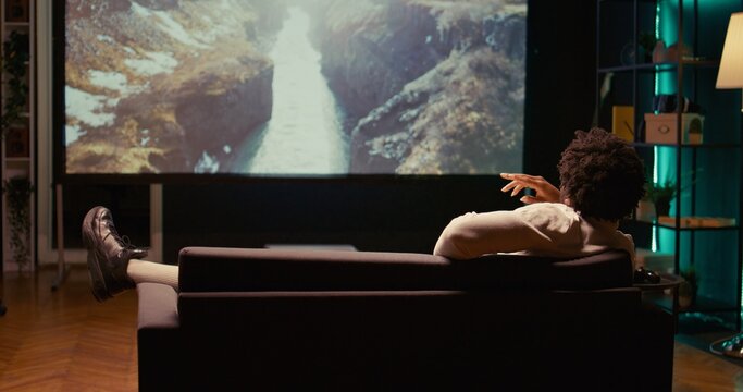 African american man watching nature documentary with beautiful cinematography on video projector. Person enjoying mountains and rivers scenery shots in home theatre, relaxing on couch