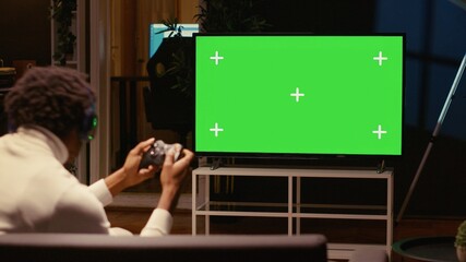Man in dimly lit apartment playing video games on green screen widescreen smart TV, enjoying day...