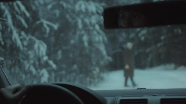 Young woman driving a car through snowy forest in the evening and stopping when creepy alien appearing on the road and walking to her. Rack focus shot