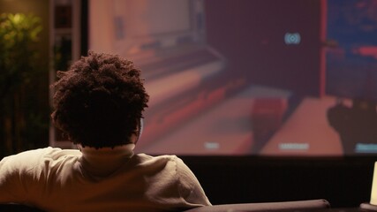 African american man watching video on widescreen smart TV of FPS in dimly lit living room. Player...