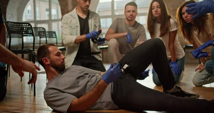 A bearded brunette man ties a medical tourniquet on her leg during a practical lesson in first aid and demonstrates it to others being trained