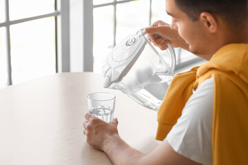 Young man pouring purified water into glass from filter jug at table in kitchen