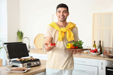 Young man with board of fresh vegetables cooking tasty sausages on modern electric grill in kitchen