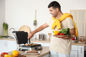 Young man with board of fresh vegetables cooking tasty sausages on modern electric grill in kitchen