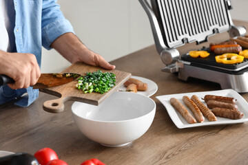Young man cooking tasty sausages on modern electric grill and making salad of fresh vegetables in kitchen