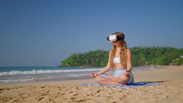 Woman in VR headset meditates on sandy beach, virtual reality mindfulness session beside sea, tech wellness trend, female practices yoga, VR mental health app, tranquil ocean backdrop. Slow motion.