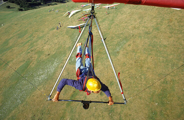 Pilot's perspective, a young man hangs in a hang glider and flies over the launch site, where other...