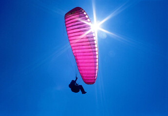 a purple, pink paraglider flies in the blue sky, backlit by the sun and lots of sunbeams
