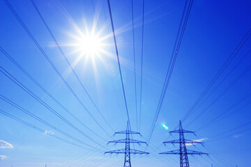 Two high-voltage pylons with high-voltage lines backlit by blue sky and sun