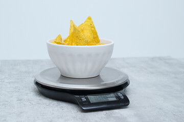 Salted tortilla nacho chips in a white bowl on a kitchen scale, grey marble table