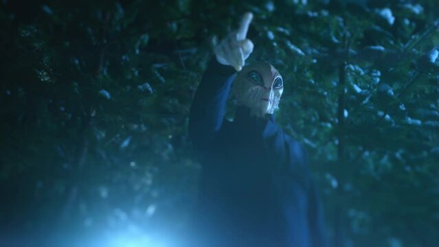 Extraterrestrial alien standing in dark forest at night, rasing one arm with long fingers and pointing at something or using superpower. Low angle view