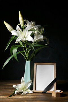 Blank funeral frame and vase with lily flowers on wooden table against dark background