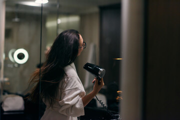 A joyful woman, with headphones in her ears, playfully dries her hair with a hairdryer in the...