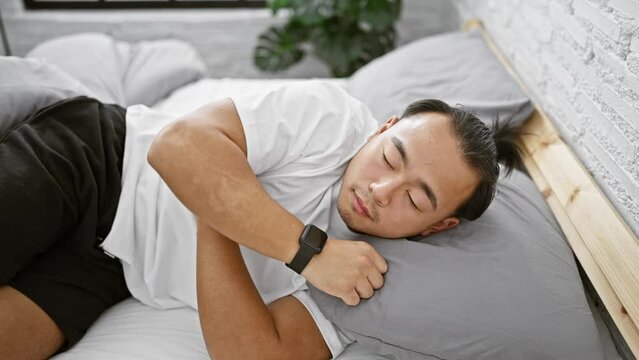 Exhausted young chinese man turning on alarm clock, surrendering to sleep in a cozy bed inside his comfy bedroom