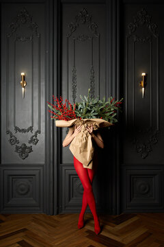 A long girl in red tights and high-heeled shoes holds a huge bouquet of fir branches and red berries wrapped in cardboard paper, she closes with a bouquet, photo studio with grey wall background