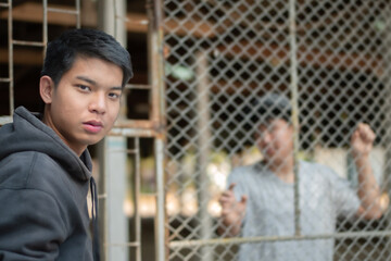 Asian preteen boy comes to visit his friend who was inside the metal fence around detention center because he was detained for inappropriate behavior, soft focus, freedom and detention concept. - obrazy, fototapety, plakaty
