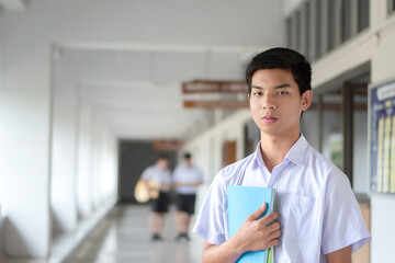 Portrait of asian secondary schoolboy holding book and standing in front of the room and waiting...