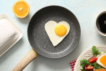 Frying pan with tasty fried egg in shape of heart, cut orange and cup of coffee on light background